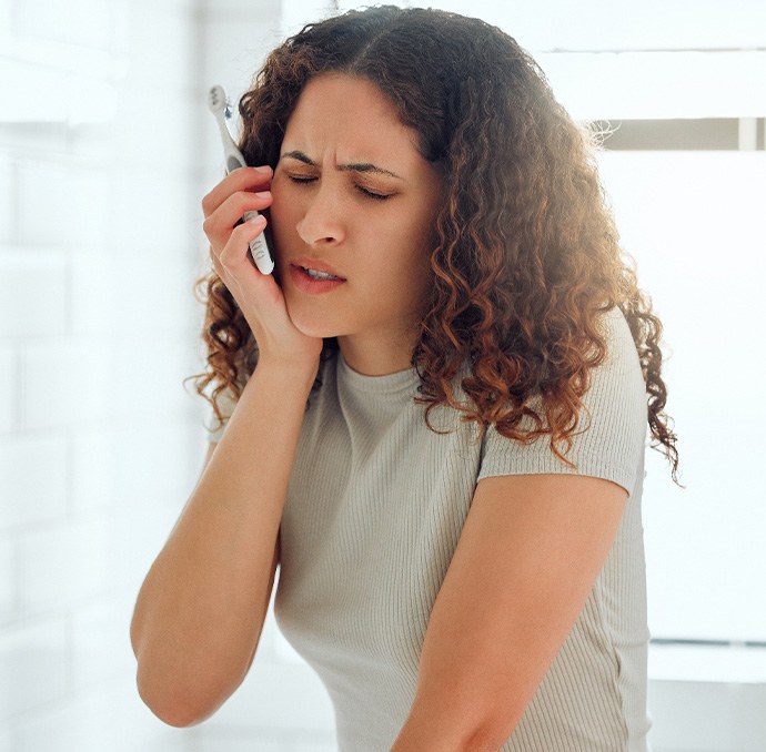 Woman with curly hair holding toothbrush and touching jaw in pain