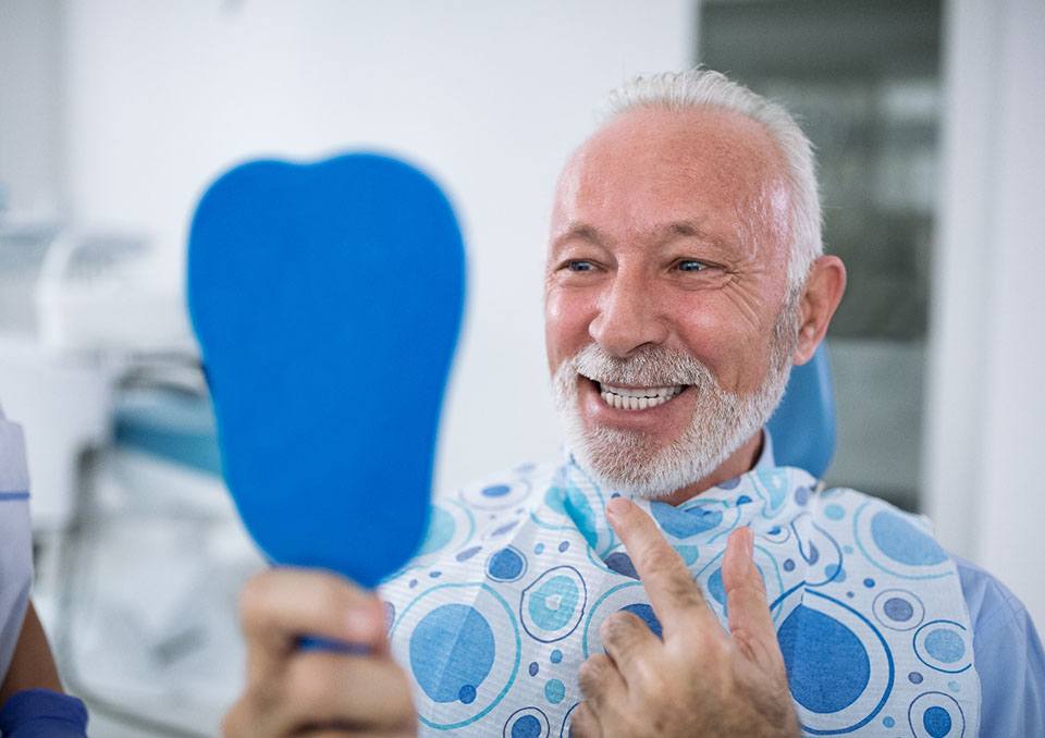 Senior man checking smile in mirror after getting implant dentures in Richardson, TX